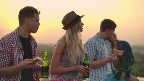 Two-couples-in-love-eat-pizza-and-drink-beer-on-the-roof.-Girls-shyly-look-at-their-boyfriends.-In-one-couple-the-girl-laid-her-head-swiftly-on-the-shoulder-of-her-young-man.